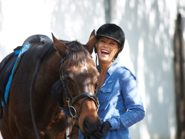 A woman in a blue shirt and black helmet smiles joyfully while standing beside a brown horse, Big Dee’s Tack and Vet Supply provides equine supplies and equipment for individual horse owners, businesses, and clinics across the United States and achieved greater customer loyalty with RFgen.